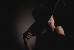Dramatic dark studio portrait of elegant woman in black wide hat and black dress. Hidden eyes.