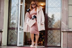 Maxi plus size rich african american model woman in dusty rose silk dress with coat and gloves, sunglasses posed outdoor near door of hotel.